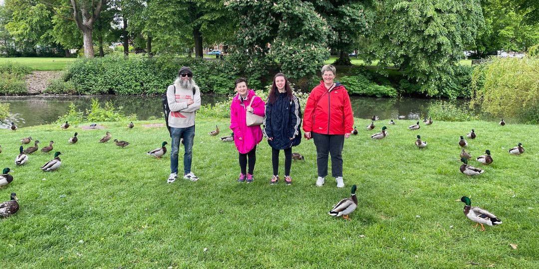 Three women and one man stand in a green park in front of a river surrounded by ducks