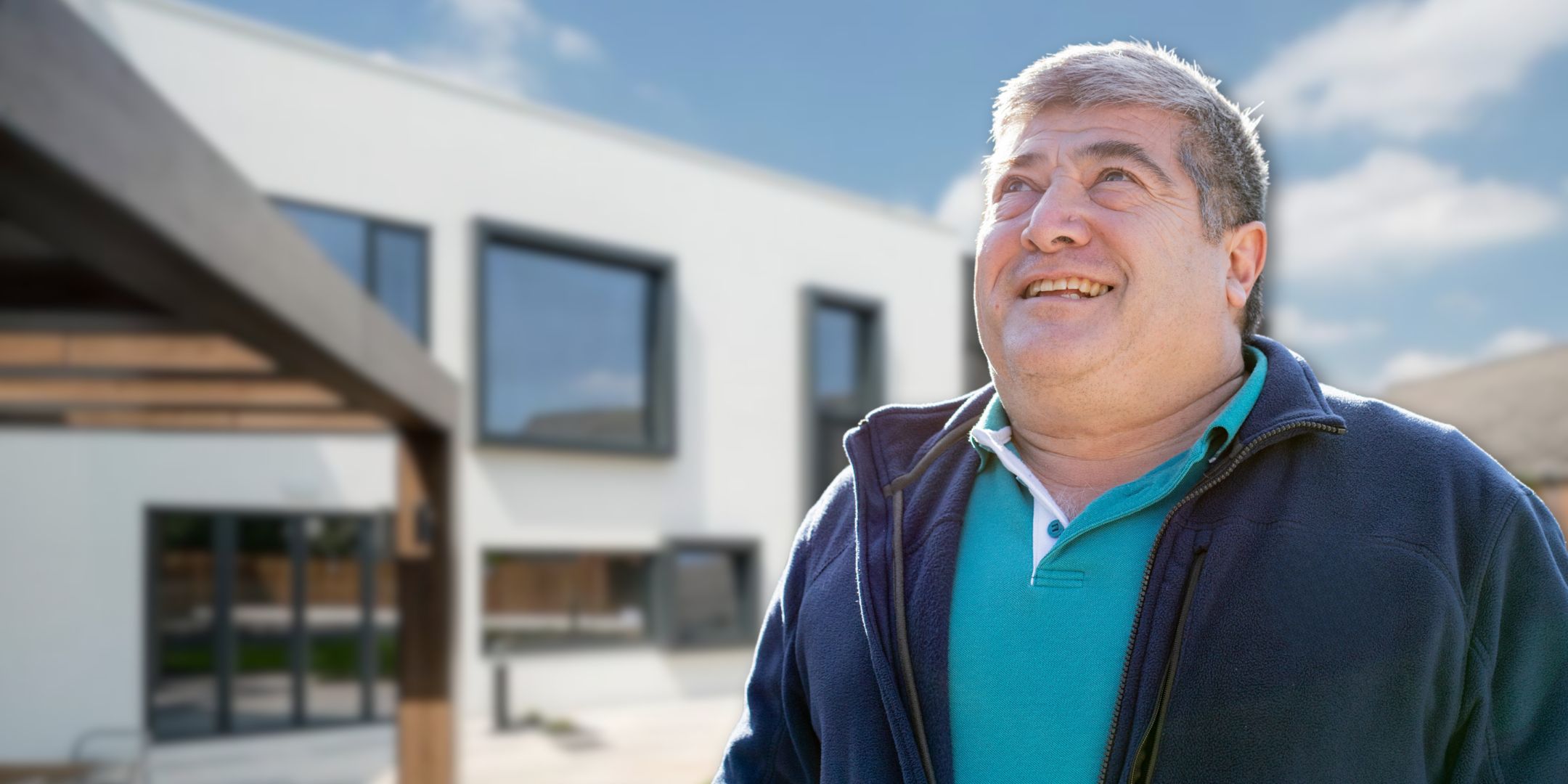 A smiling white man in a blue polo and navy fleece stands in front of a building on a sunny day.