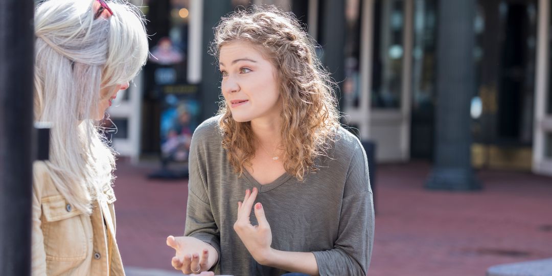 A lady with shoulder length blonde curly hair wearing a green shirt talking to a lady with mid length grey hair