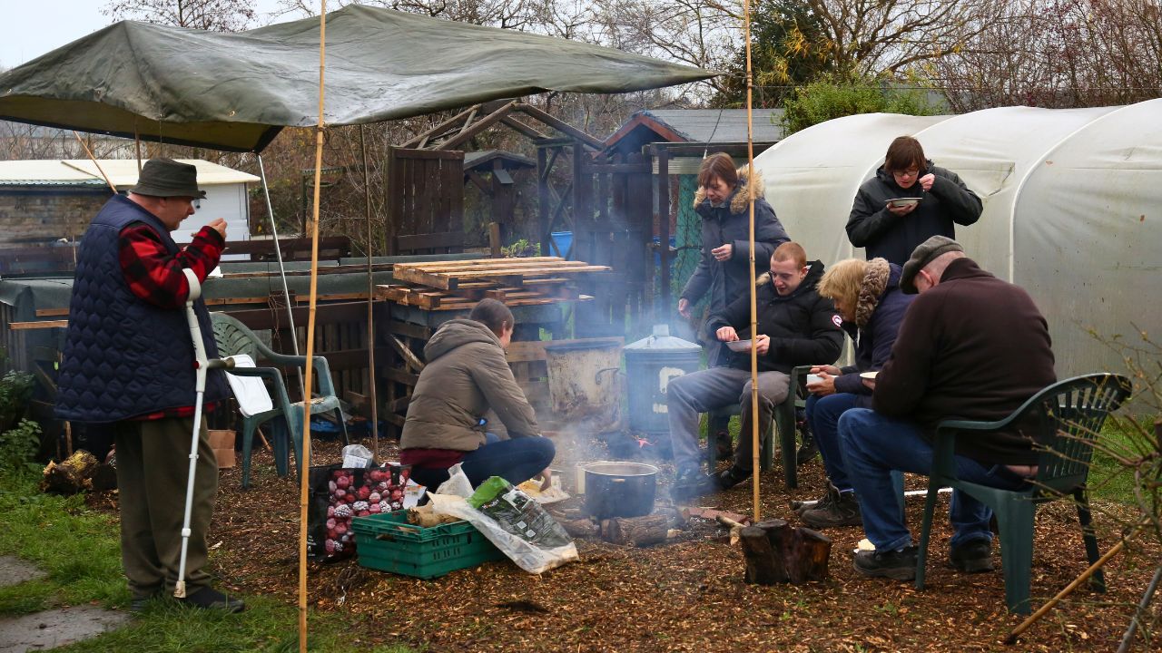 Group of people eating soup around a fire outdoors.